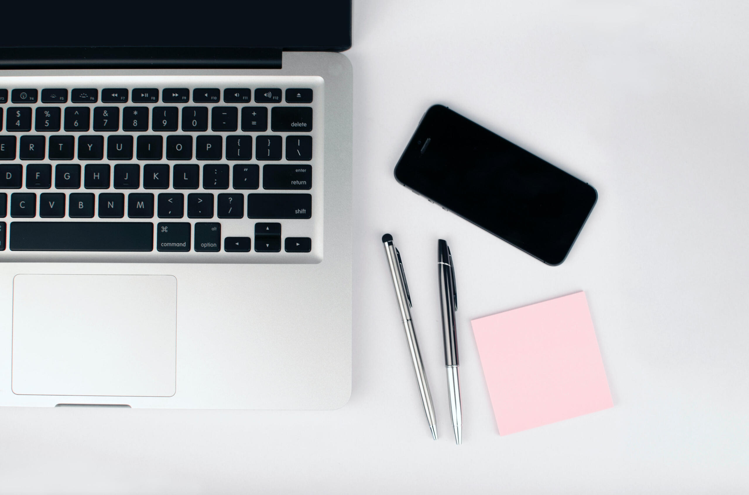 Aerial photo of a Macbook laptop on a table next to an iPhone, two pens and a pink Post-It note pad